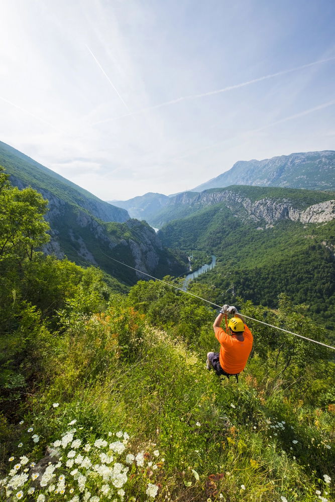 An individual zip-lining over a lush green valley with a winding river, surrounded by majestic mountains under a clear sky in Omis.