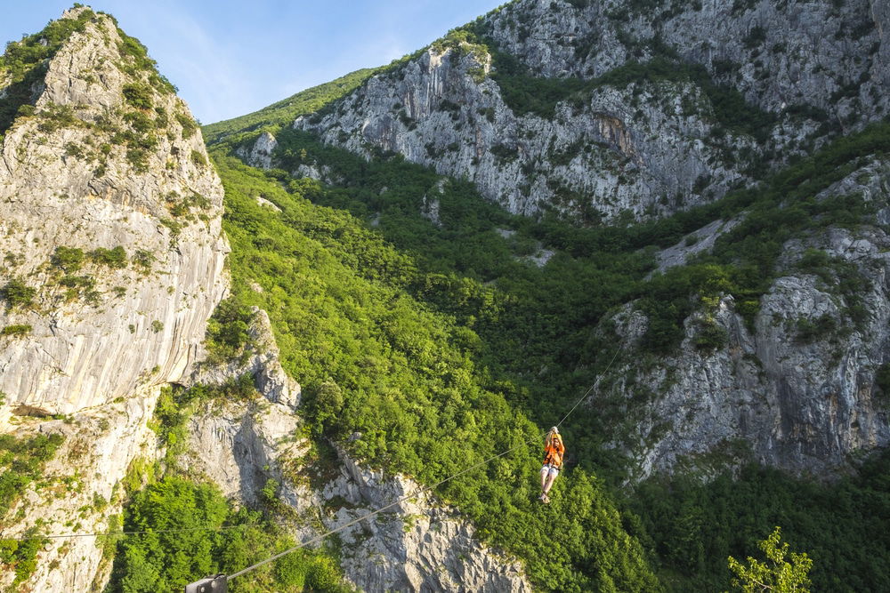 A person in orange and yellow gear is walking a tightrope stretched across a mountainous valley with lush green vegetation below, similar to the adventurous spirit you'd find ziplining in Omis.