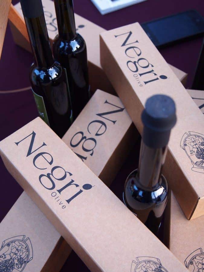 Several cardboard boxes with "Negri Olive" branding lie scattered, partially covering a few dark glass bottles with black caps. The background is a dark maroon surface.