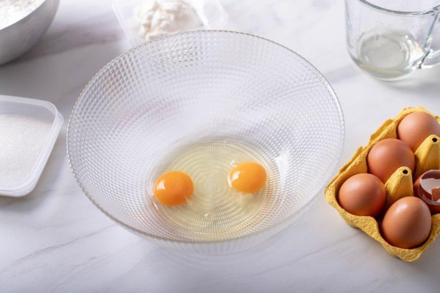 Two eggs in a bowl next to other ingredients for making Croatian Kroštule, sweet pastry knots.