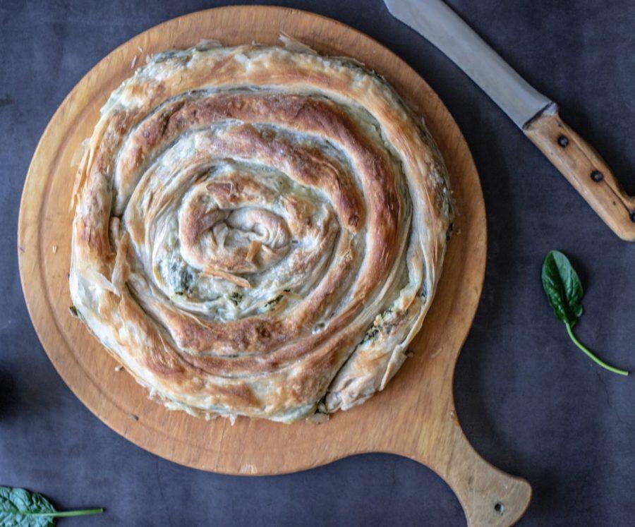 A round, spiral-shaped pastry on a wooden cutting board with a knife and spinach leaves beside it.