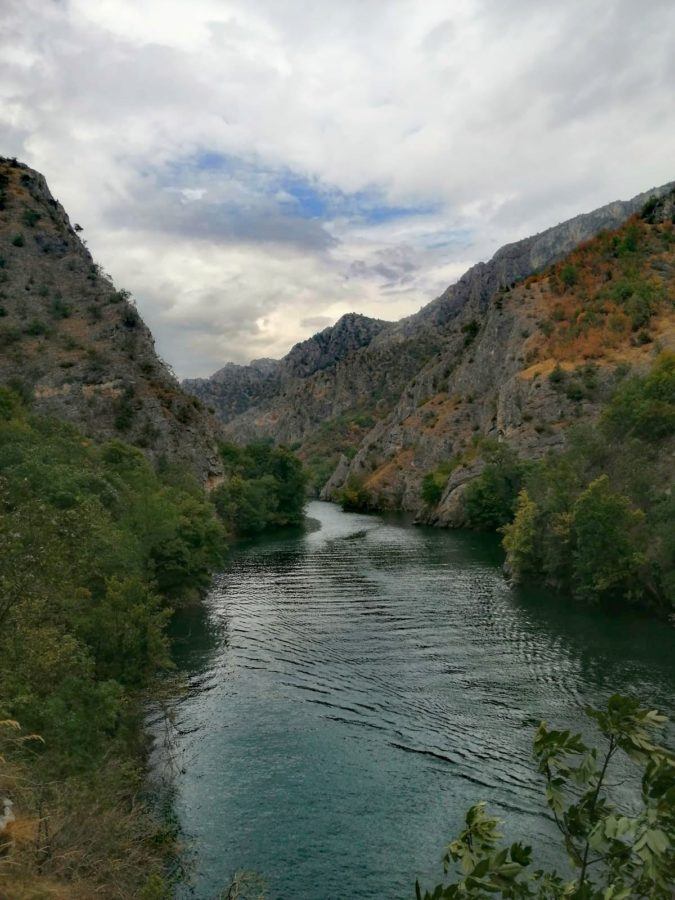 A winding river flows through a mountainous landscape with cloudy skies overhead. Rocks and greenery are present on either side of the river.