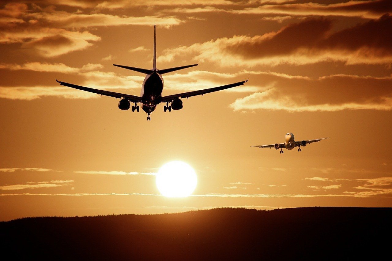 Two airplanes are flying towards the camera with the setting sun and clouds in the background. The larger plane is closer to the viewer, while the smaller one is further away.