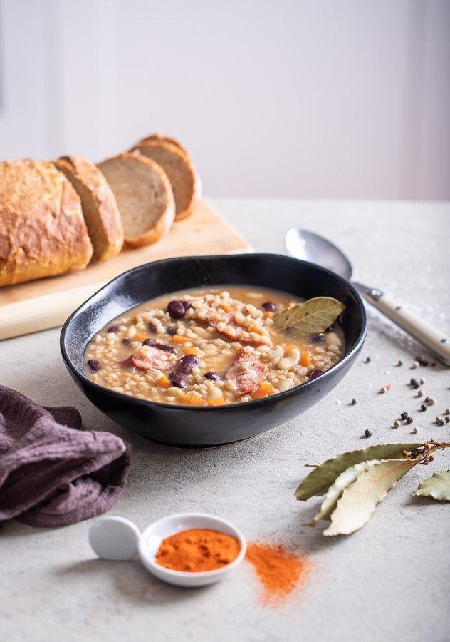 A bowl of Croatian soup with beans, barley stew, and spices served alongside bread on a table.