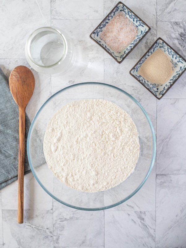 Using a wooden spoon to make somun bread, a bowl of flour sits on a marble table.