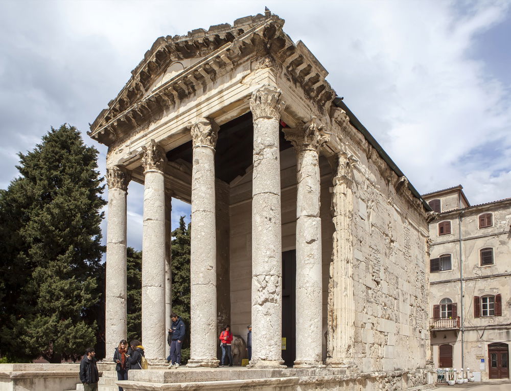 A group of people stand near the entrance of an ancient Roman temple - Temple Of Augustus with tall Corinthian columns and a partially ruined pediment, one of the must-see attractions when exploring things to do in Pula.