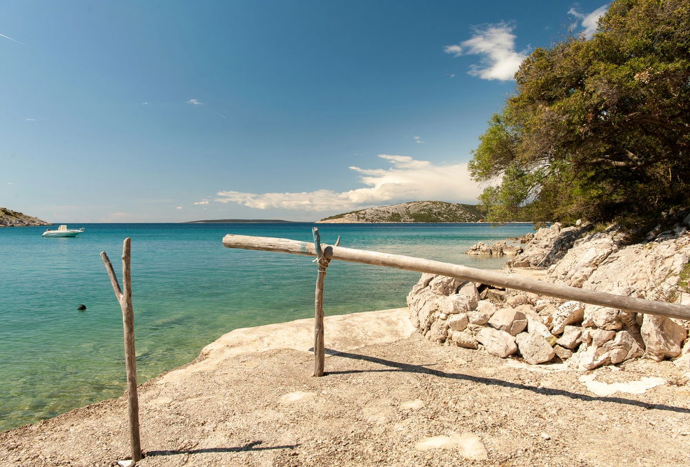 Ustrine Cover with clear blue water, a distant boat, rocky shoreline, and wooden poles in the foreground under a clear sky, highlights just some of the things to do on Cres.