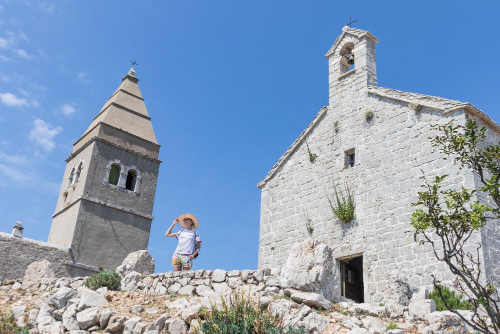 A person wearing a hat stands in front of an old stone church with a bell tower on a bright sunny day, showcasing one of the popular things to do on Cres. Accient village of Lubenice.