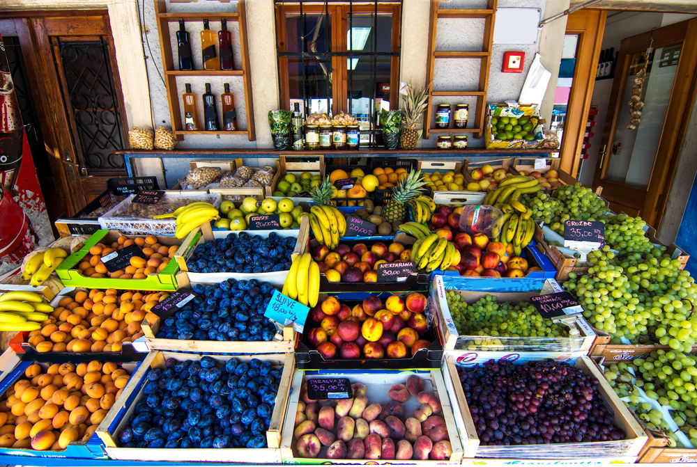 A market stall displaying a variety of fresh fruits including apricots, plums, bananas, peaches, grapes, apples, and pears, along with bottles of olive oil and jars of honey on the shelves behind. It's one of the delightful things to do on Cres.