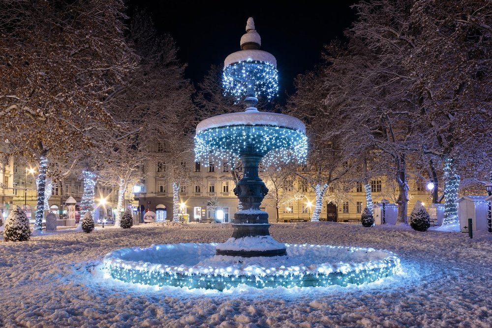 The snow-covered Zrinjevac Fountain with blue and white holiday lights on a winter night in Croatia.