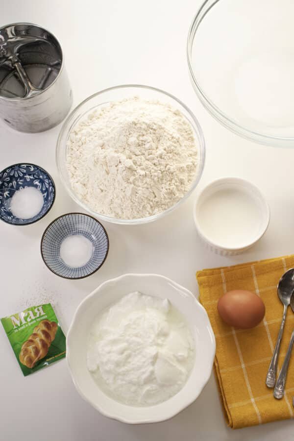 A Bulgarian recipe for mekitsi on a white table with bowls of flour, eggs, and other ingredients.