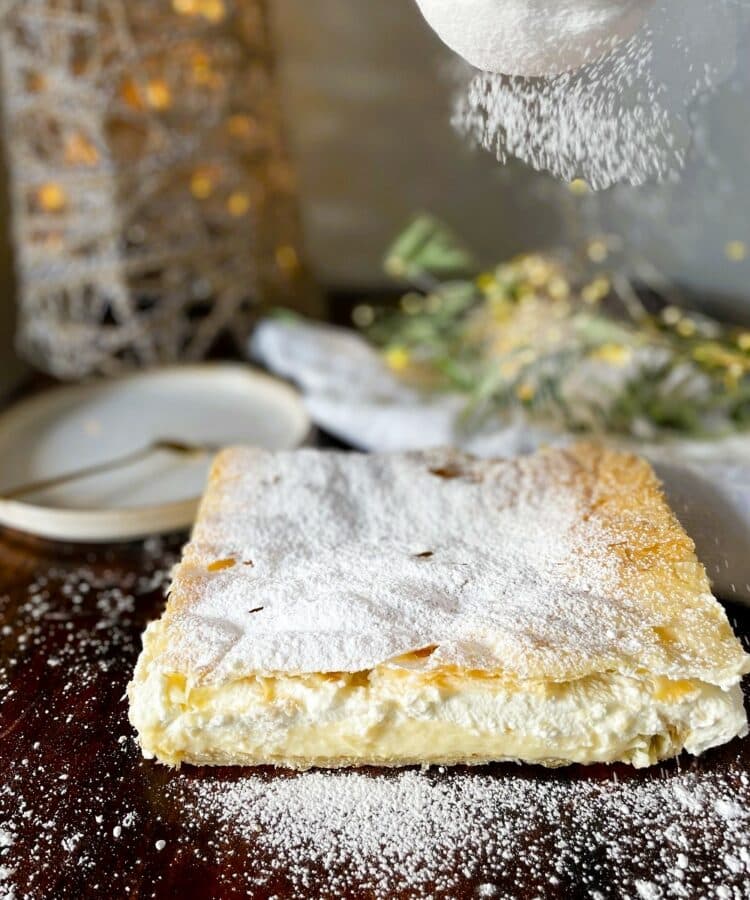 A square pastry topped with powdered sugar is placed on a dark wooden surface, reminiscent of a traditional kremšnite recipe. A small white plate and fork are in the background.