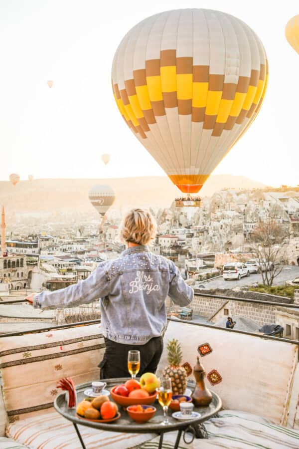 A person stands on a rooftop in Cappadocia, gazing at hot air balloons over a cityscape at sunrise, with a table set with fruits and drinks in the foreground.