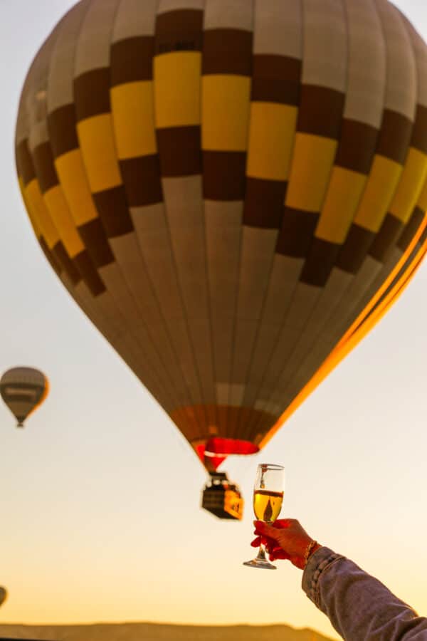 A hand holding a champagne glass with hot air balloons floating in the background during a golden sunrise in Cappadocia.