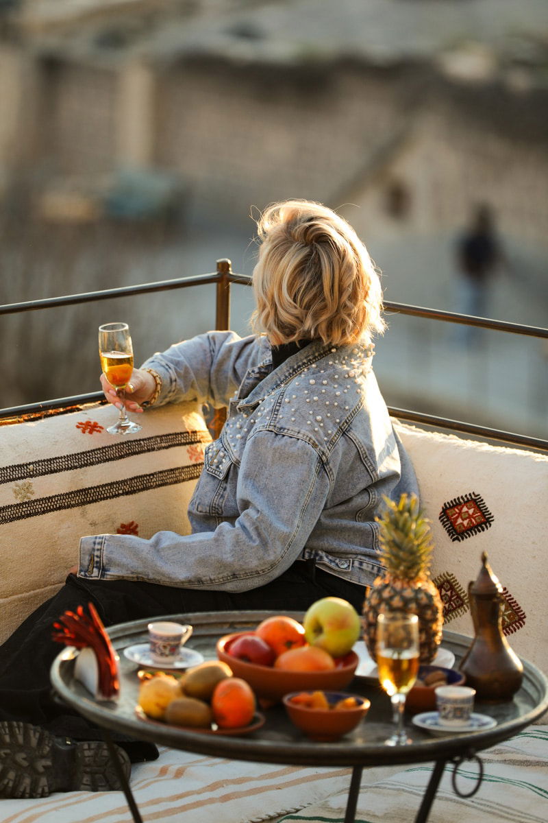 SJ in denim jacket seated on a balcony holding a glass of wine, with a table of fruits and snacks beside her at sunrise in Cappadocia.
