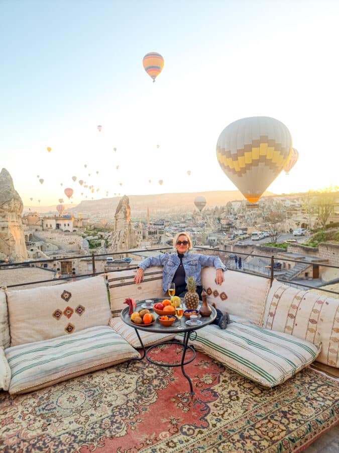 A person sitting on a cushioned bench with a breakfast spread, enjoying the Cappadocia sunrise as numerous hot air balloons rise over a rocky landscape.