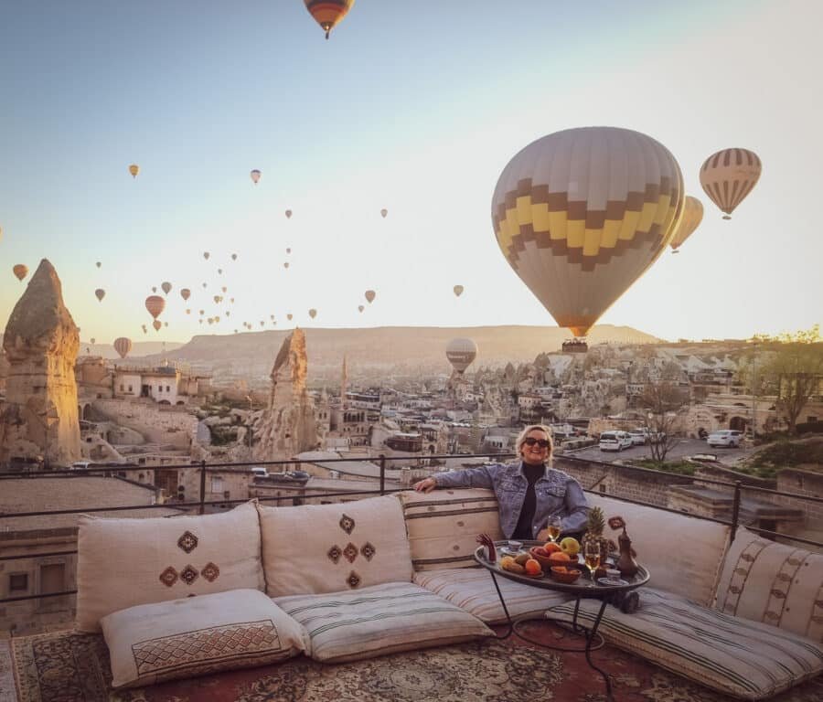 A person lounging on an outdoor sofa, smiling, with many hot air balloons floating in the sky during a Cappadocia sunrise.