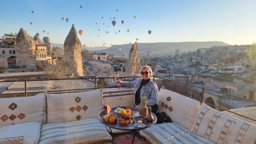 A woman stands on a terrace with a breakfast spread, overlooking a sunrise landscape dotted with hot air balloons and unique rock formations in Cappadocia, Turkey.