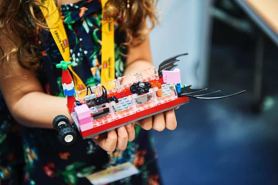 A child in Istanbul proudly holds a colorful LEGO creation featuring small wheels, transparent pieces, and dragon wings, all while wearing a yellow lanyard around their neck. This joyful moment captures the essence of family fun and playful imagination among kids.