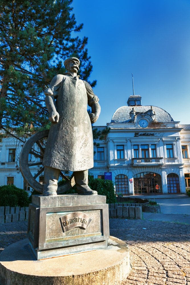 A statue of a man standing in front of a building in Kragujevac, Serbia Stara Livnica district. Wonderful building with old clock and statue of worker.