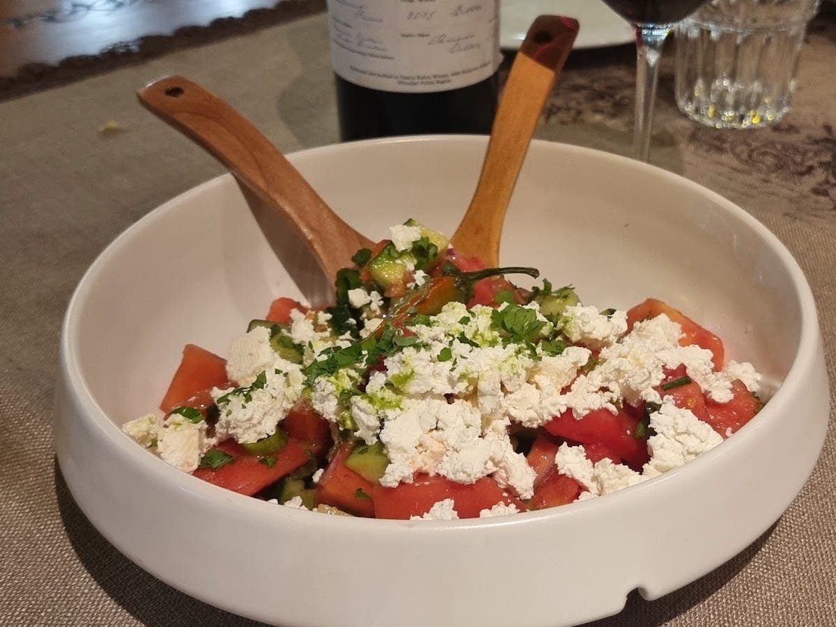 A Bulgarian salad with tomatoes and feta in it next to a bottle of wine. Shopska Salata