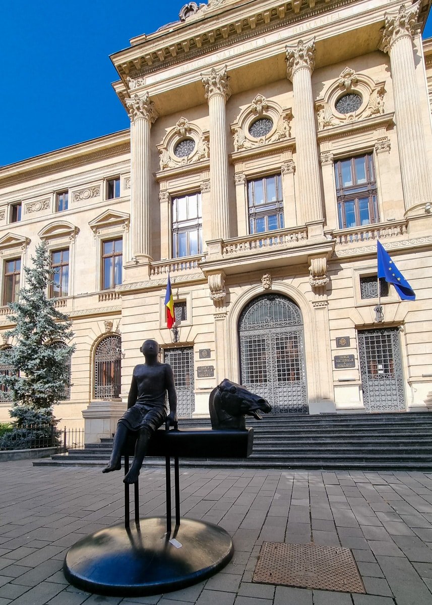 A statue of a man sitting on a bench in front of a building on a walk on Old Town Bucharest.