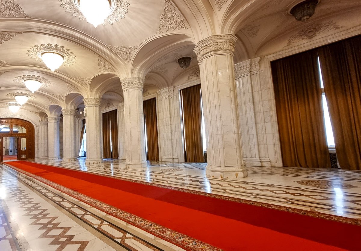 An ornate hallway with a red carpet and pillars A tapestry with at the Palace of Parliament in Bucharest.