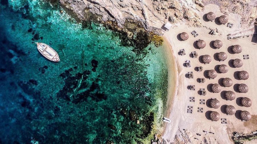 A summer aerial view of a beach in Mykonos adorned with colorful umbrellas and featuring a boat sailing in the sea.
