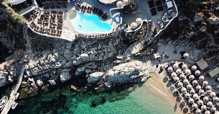 A tranquil summer aerial view of a swimming pool on a cliff overlooking the beach clubs in Mykonos.