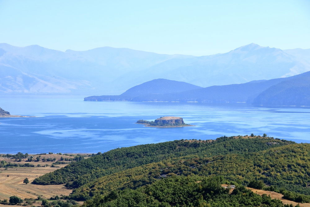 A panoramic view of a mountainous lakescape in one of the national parks in Albania - Albania - the Prespa National Park- Lake Prespa featuring a small island in the center, surrounded by rolling green hills under a clear blue sky.