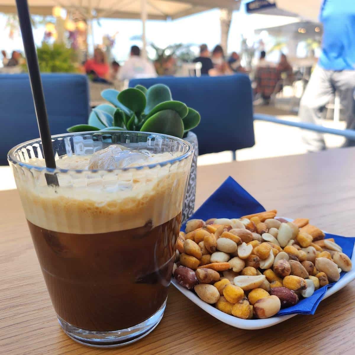 A glass of iced coffee sits on a table next to a plate of nuts in Greece.