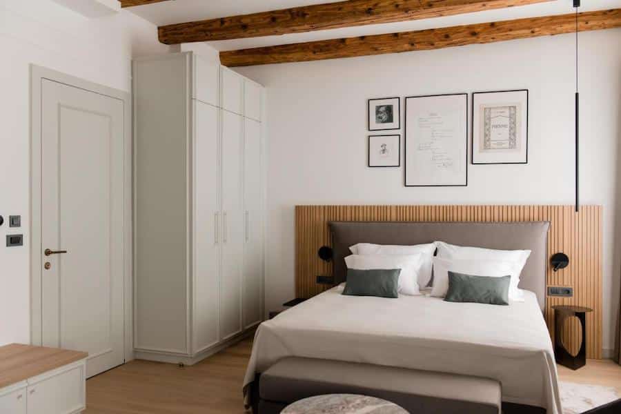 A minimalist bedroom, perfect for those seeking where to stay in Zagreb, features a neatly made bed with white linens and green cushions, a white wardrobe, wooden ceiling beams, framed art on the wall, and a small bench at the foot of the bed.