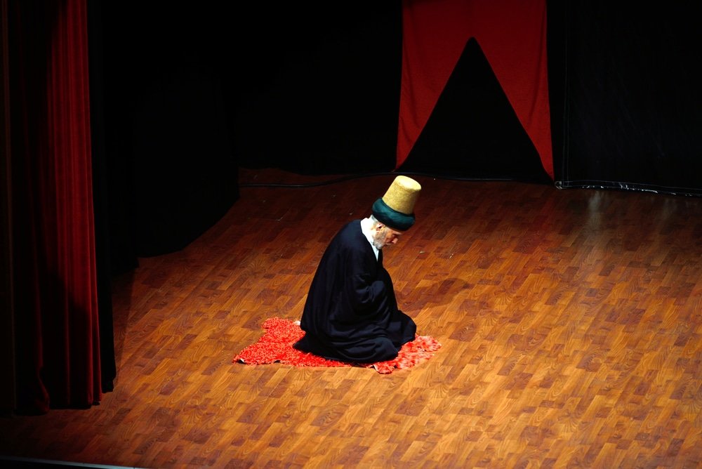 A man in a black robe kneels on a wooden floor - the Whirling Dervishes in Konya.