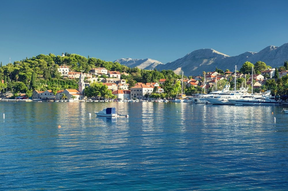 Coastal town in Croatia with Mediterranean architecture by a calm blue sea, yachts moored near tree-covered hills under clear skies.