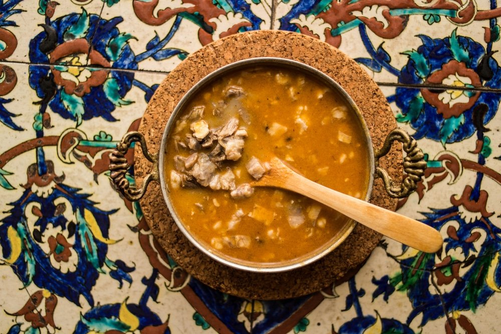 A bowl of Kelle Paça Çorbası soup with a wooden spoon on a tiled floor, inspired by Turkey.