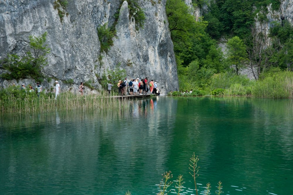 Group of visitors on a wooden walkway by a turquoise lake with a rocky cliff in the background, illustrating why Plitvice Lakes is worth visiting.