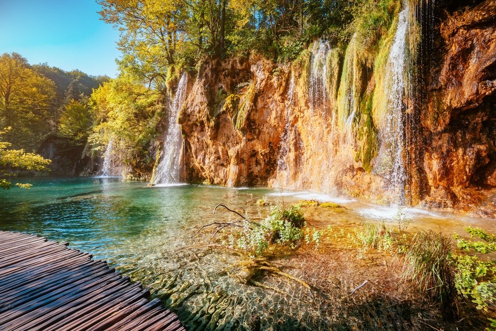 A scenic view of Plitvice Lakes, featuring waterfalls with lush vegetation alongside a wooden boardwalk.