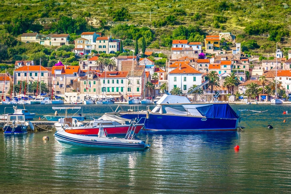 Boats moored in a serene harbor with a colorful hillside village in the background, reminiscent of Croatia.