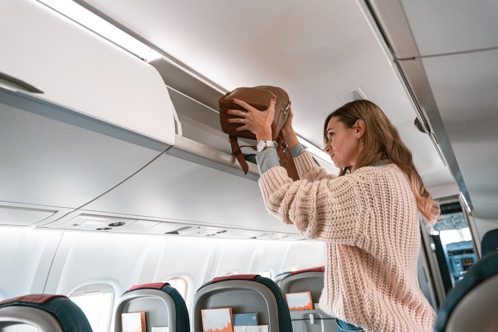 Woman placing a carry-on bag into an overhead compartment on an airplane, dressed in a comfy travel outfit.
