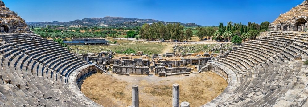 Panoramic view of an ancient Roman amphitheater in Aydın Province, with surrounding ruins and a scenic backdrop of rolling hills under a clear sky.