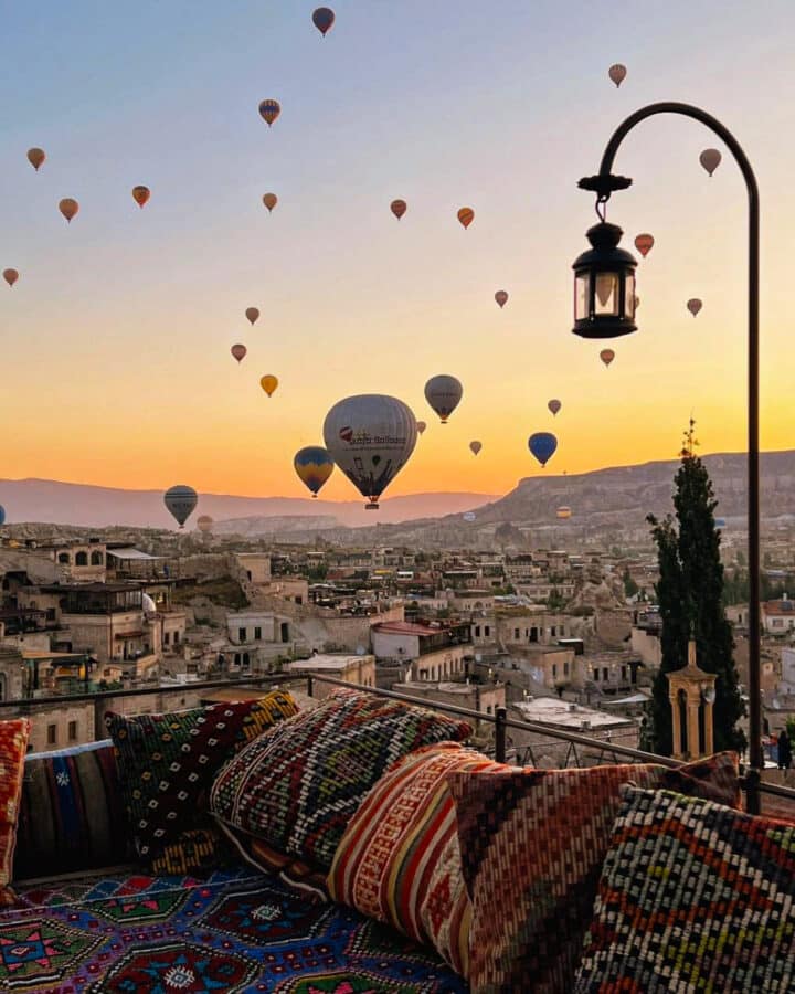 Hot air balloons float over a historic town at sunrise, viewed from a terrace adorned with colorful cushions.