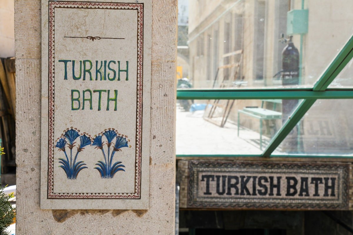 Signs labeled "turkish bath" with floral designs, attached to a stone wall in Goreme, Cappadocia, on a sunny city street, with blurred background of a building.
