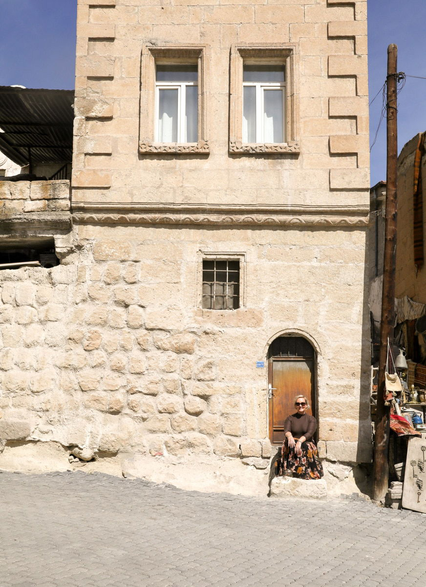SJ sits beside a stone building with a wooden door in Goreme, under the shadow of an adjacent building on a sunny day.
