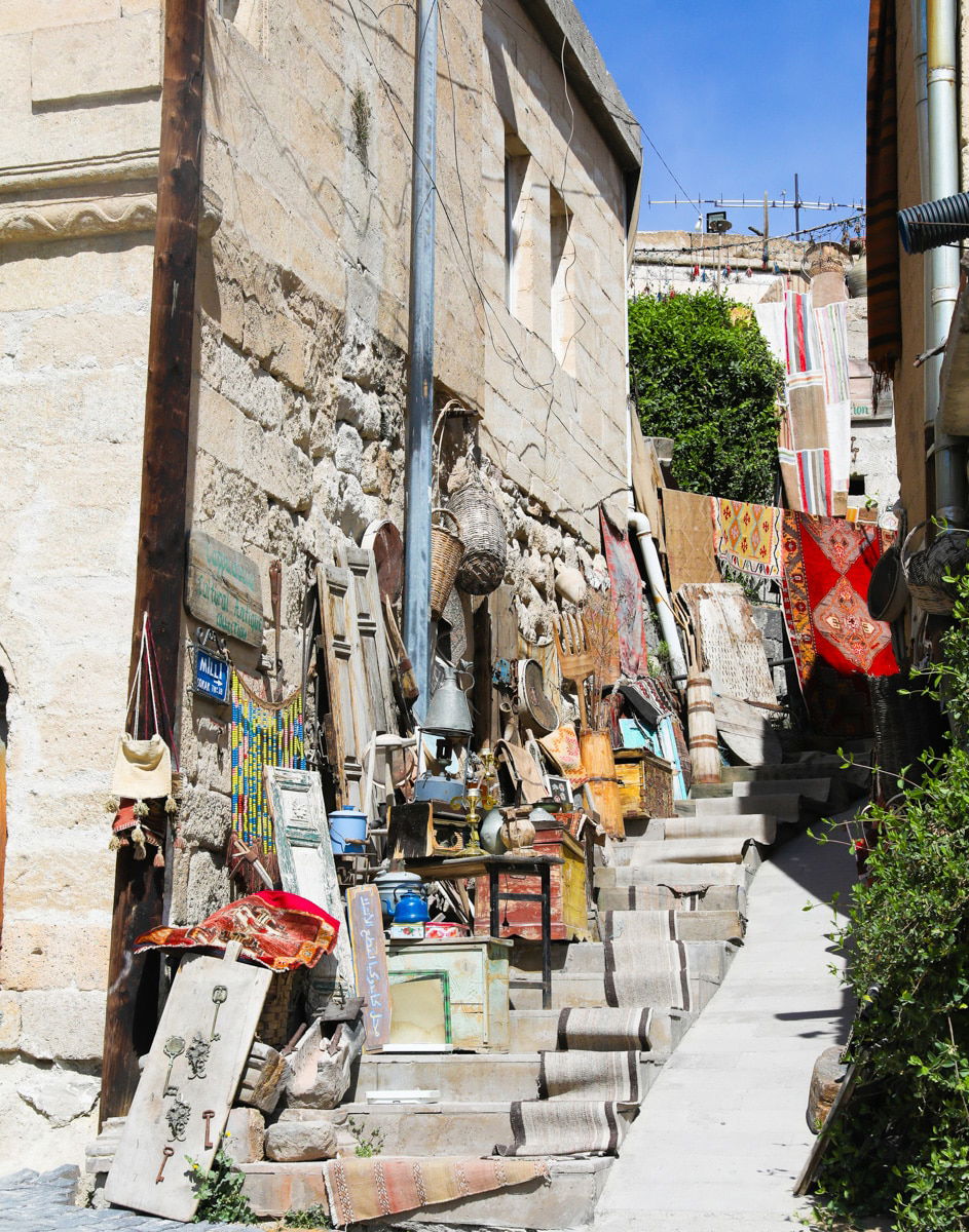 Narrow stone stairway between buildings with various rugs and artworks displayed for sale in a sunlit market in Goreme, Cappadocia.