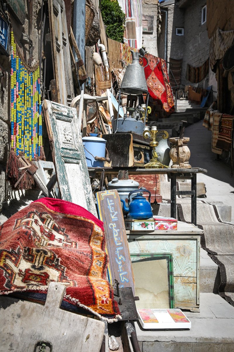 A variety of antique items and colorful textiles displayed on a rustic street market in Goreme, Cappadocia, including carpets, mirrors, and old metallic objects.
