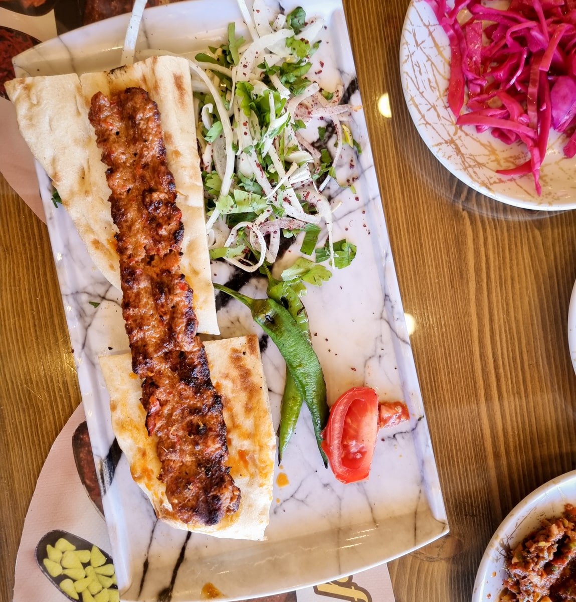 A plate of adana kebab served in Goreme, Cappadocia, with flatbread, grilled green pepper, and tomato, accompanied by sides of onion salad and pickled cabbage.