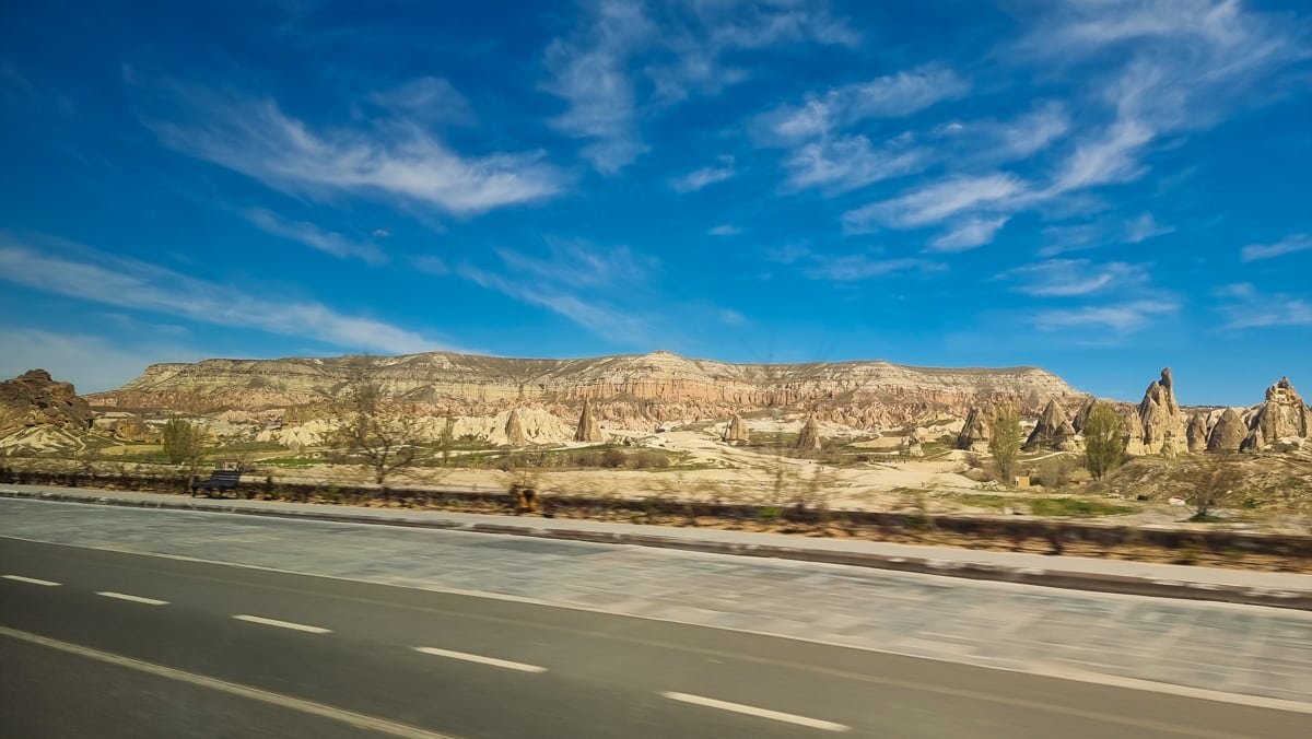 A scenic view of a rugged landscape with unique rock formations in Goreme under a vast blue sky with streaky clouds, taken from a roadside perspective.
