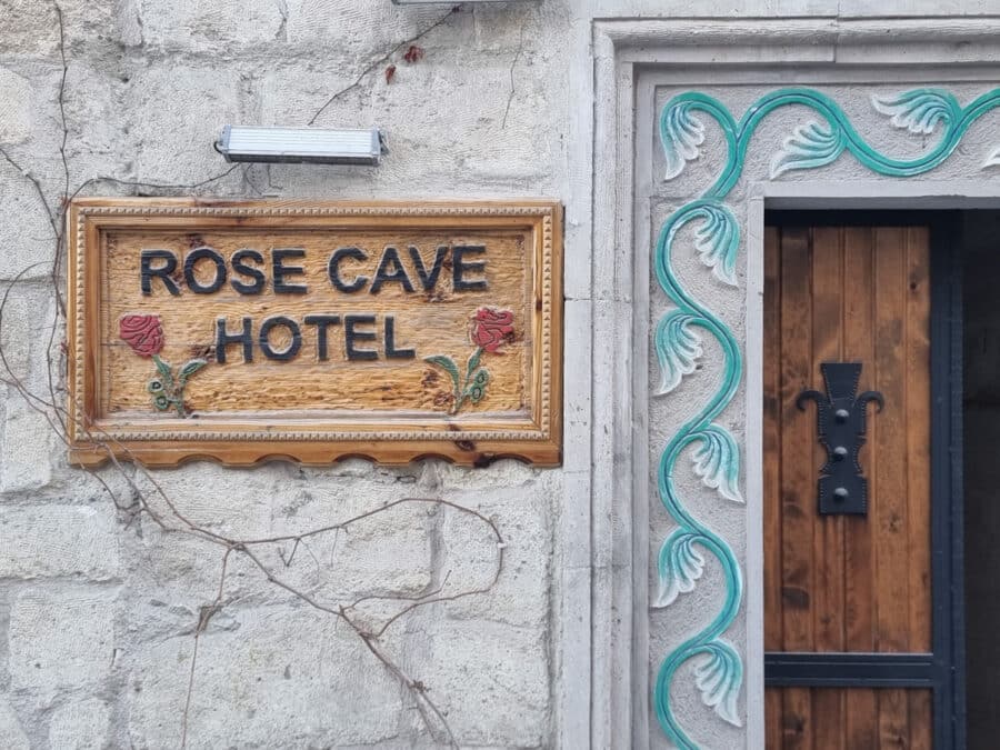 Carved wooden sign reading "Rose Cave Hotel" in Goreme, on a stone wall next to a wooden door with ornate teal decorations.