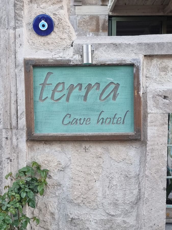 A weathered wooden sign that reads "Cappadocia Terra Cave Hotel" mounted on a stone wall, with an eye-shaped amulet above it.