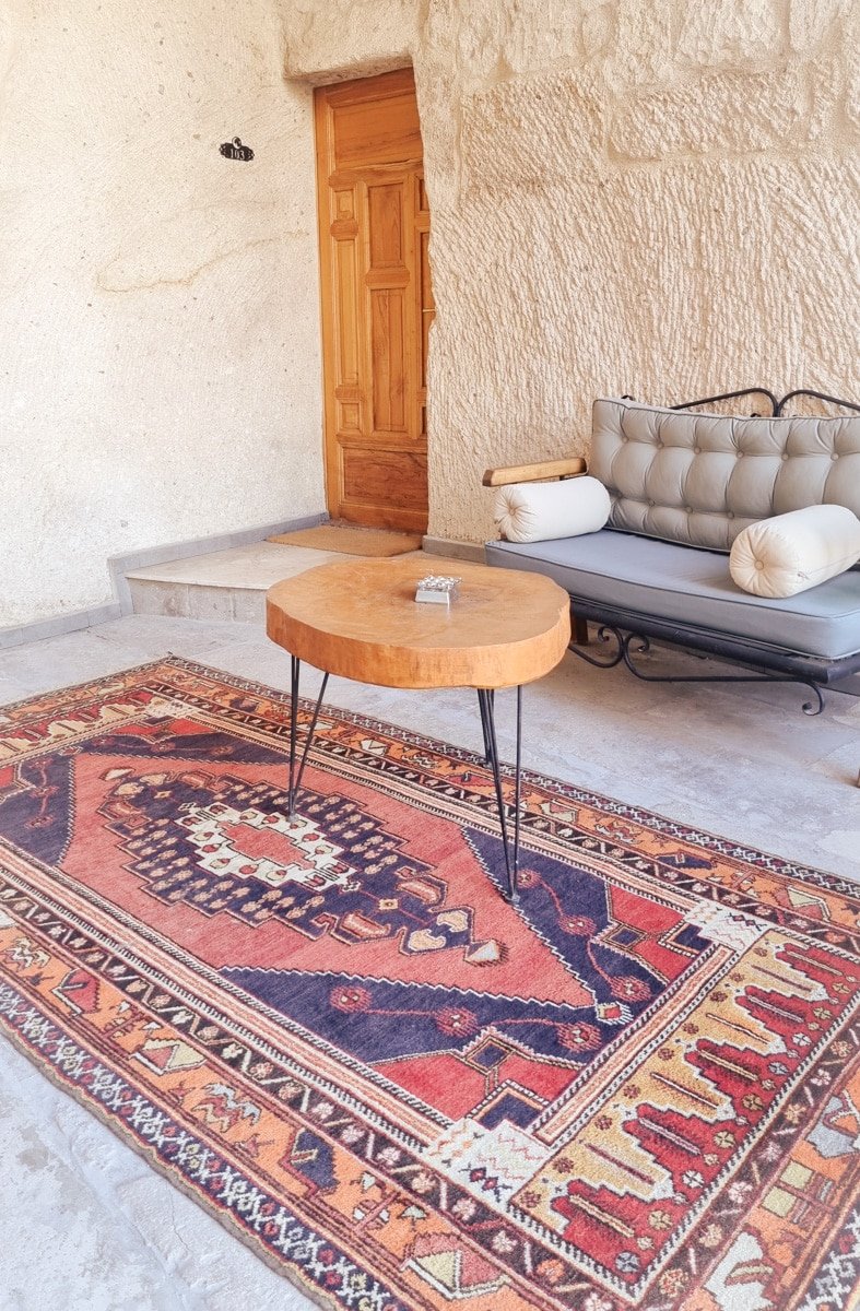 A rustic outdoor sitting area in Goreme, Cappadocia, with a wooden coffee table on an ornate rug, a beige sofa, and a wooden door in a stone wall.